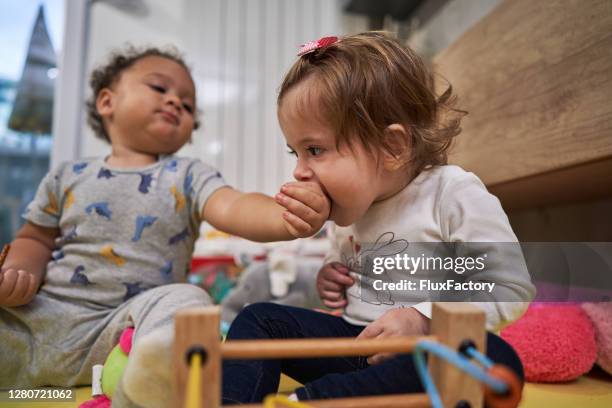 nieuwsgierig babymeisje dat gemengde hand van de rasbabyjongen bijt, terwijl zij problemen met primaire tandgroei heeft - bijten stockfoto's en -beelden