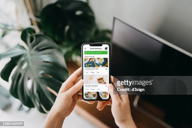 close up of young asian woman's hand using meal delivery service and ordering food online with smartphone in the living room at home, against green potted plants - smartphone stockfoto's en -beelden