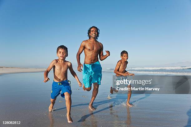 young indian man and boy's (7, 11, 25 years old) running along waters edge, muizenberg beach, cape town, western cape province, south africa - indian family vacation stock pictures, royalty-free photos & images