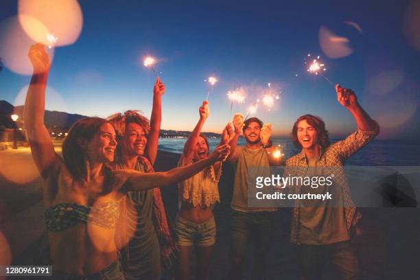 gruppe von freunden spielen mit wunderkerzen und feuerwerk am strand bei sonnenuntergang. - new years eve 2019 stock-fotos und bilder