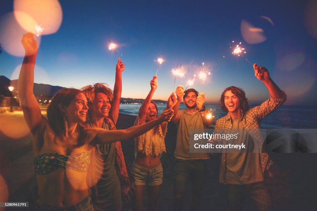 Group of friends playing with sparklers and fireworks on the beach at sunset.