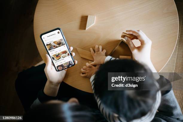 overhead view of young asian mother using meal delivery service and ordering food online with smartphone while spending time playing wooden puzzle with little daughter in the living room at home - family ordering at restaurant stock pictures, royalty-free photos & images