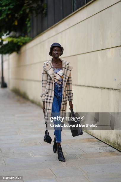 Model wears a bob hat with leopard print inner lining, a beige and black checked trench coat, a colored top with printed patterns, blue cropped denim...