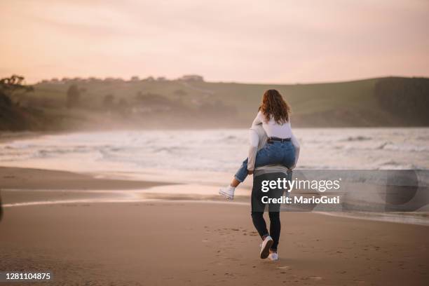 rückansicht eines jungen paares, das bei sonnenuntergang an einem strand spazieren geht - person auf den schultern tragen stock-fotos und bilder