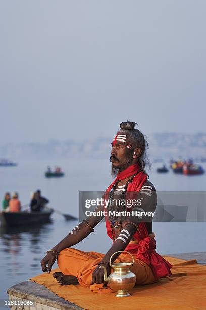 sadhu or holy man performing manas puja. this religious ritual takes place during sunrise. the little brass pot he uses is called a kamandal. buddhist tourists in boat watching ritual. ganges river, varanasi, india. (mr) - sadhu stock-fotos und bilder