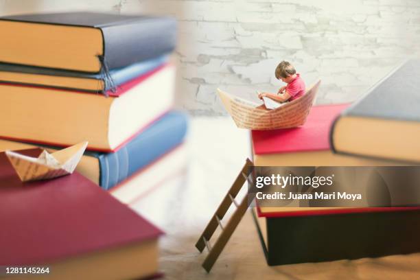 a boy reading, inside a little paper boat. on top of a pile of giant books - literatura fotografías e imágenes de stock