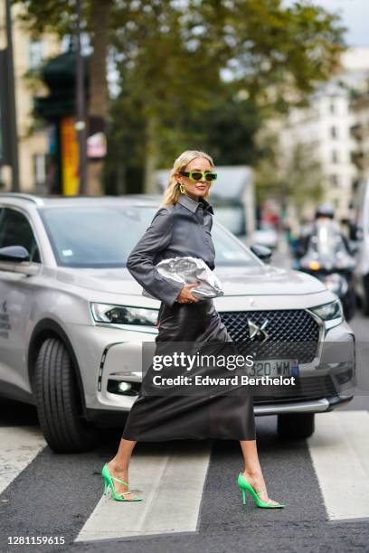 Leonie Hanne wears green sunglasses, earrings, a gray leather shirt, a silver shiny Bottega Veneta Pouch bag, a black leather skirt, green high heels...