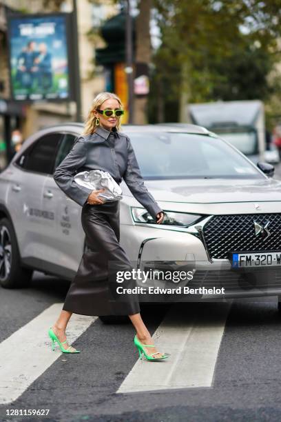 Leonie Hanne wears green sunglasses, earrings, a gray leather shirt, a silver shiny Bottega Veneta Pouch bag, a black leather skirt, green high heels...