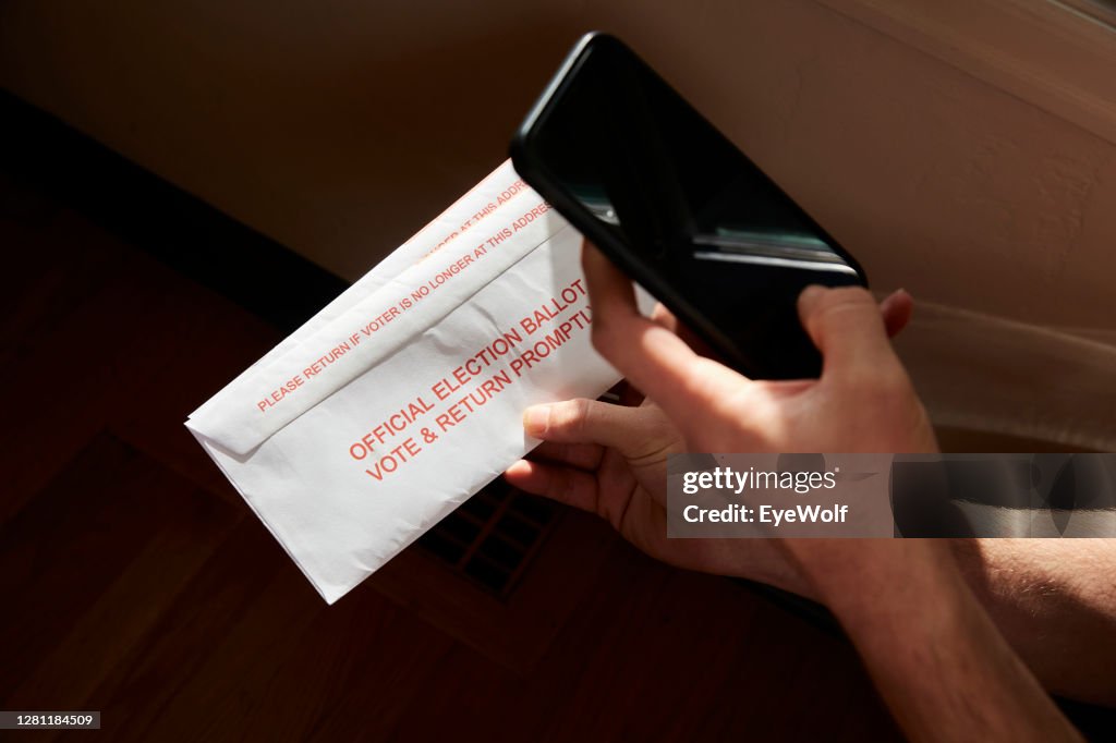 A person taking a photo of their election ballot envelop inside their home, with a rainbow streaming across it.