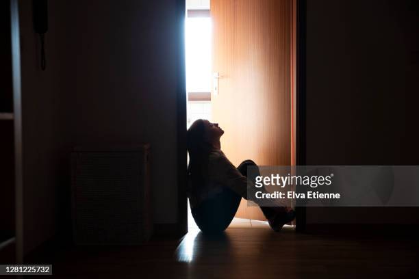 backlit teenager sitting in a dark indoor doorway in contemplation - kindertijd stockfoto's en -beelden