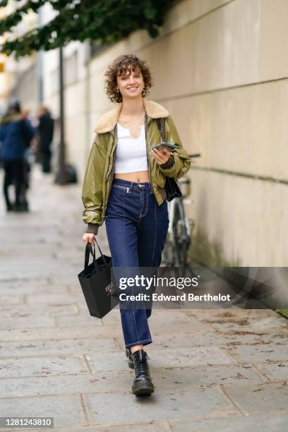 Model wears a green khaki aviator / bomber shiny jacket, a white crop top, blue cropped denim jeans pants, black leather boots, outside Kenzo, during...