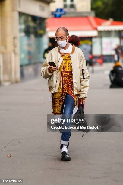 Guest wears a beige bomber jacket, a long shirt with printed poster, blue hems denim jeans, white socks, black leather shoes, glasses, a white face...