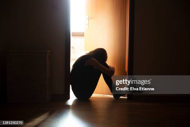 despairing teenager sitting with head on knees in the dark frame of a doorway, backlit by a room behind flooded with daylight - alleen één tienermeisje stockfoto's en -beelden