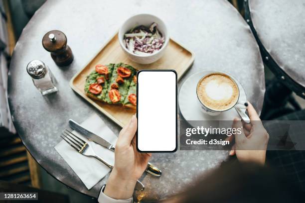 overhead view of young asian woman enjoying a healthy light meal, sourdough toast with smashed avocado and a cup of coffee in cafe. enjoying a relaxing afternoon. she is holding a frameless smartphone with blank screen for mockup - afternoon stock pictures, royalty-free photos & images