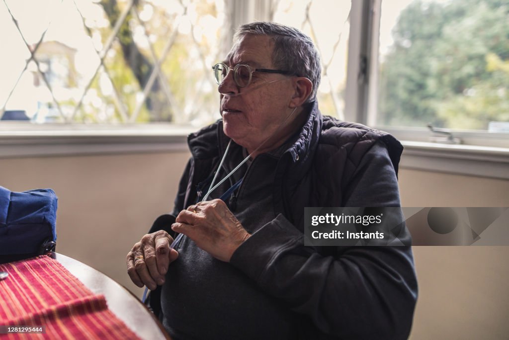 Em uma cozinha, um idoso está ajustando o tubo de seus óculos de oxigênio antes de tomar café da manhã.
