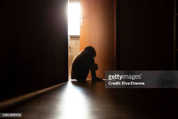 despairing child sitting with head on knees in the dark frame of a doorway, backlit by a room behind flooded with daylight - solitario foto e immagini stock