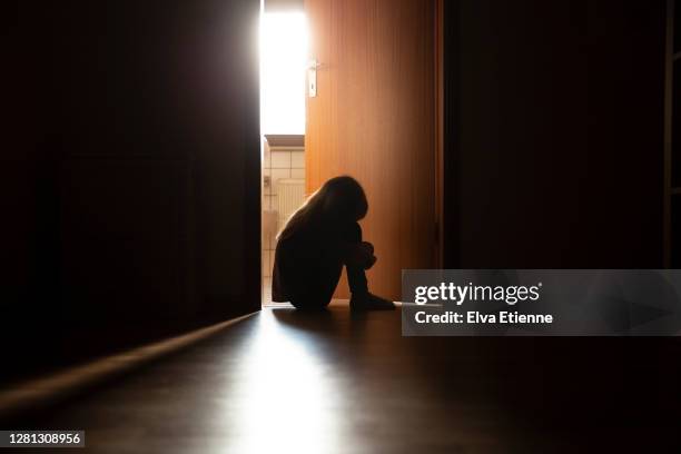 despairing child sitting with head on knees in the dark frame of a doorway, backlit by a room behind flooded with daylight - sombre bildbanksfoton och bilder