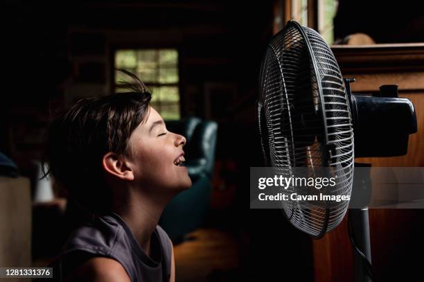 young boy with eyes closed cooling off in front of a fan in dark room. - boy in wind stock-fotos und bilder