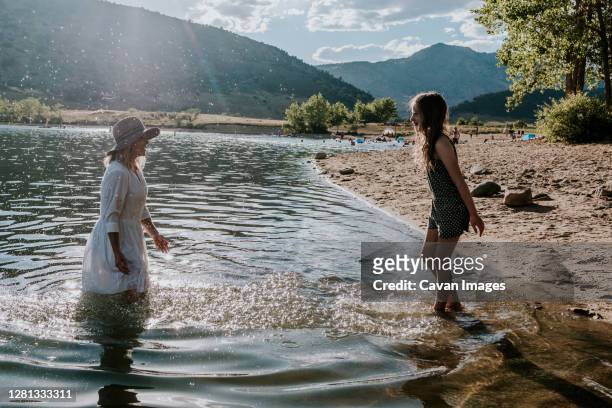mom and daughter splashing and playing on the shore of a lake - girls in wet dresses stock-fotos und bilder