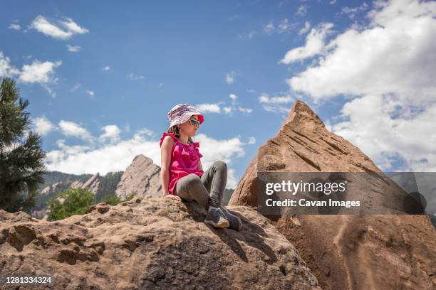 young girl with hat sits on rock with mountains behind after hiking - boulder rock stock-fotos und bilder