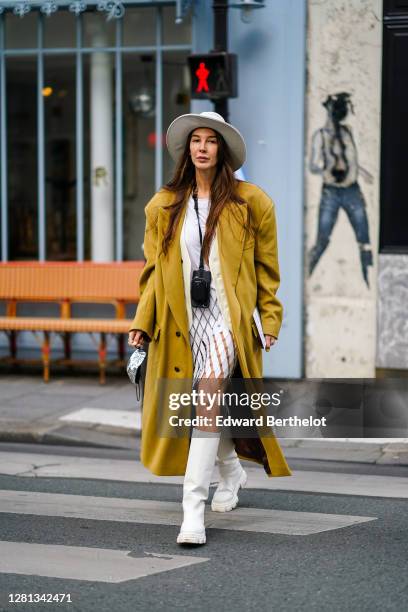 Estelle Chemouny wears a white hat, a mustard-yellow long oversized coat, a white jacket, a Prada small bag, a white and clear mesh dress with argyle...