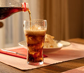 Pouring Cola from Bottle into Glass and Fizz with Ice Cubes on Table Against Blurred Livinroom  Background