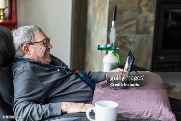 hombre mayor relajándose y pasándoselo bien mientras está sentado en un reclinable y viendo contenido divertido en su tableta. - reality tv fotografías e imágenes de stock