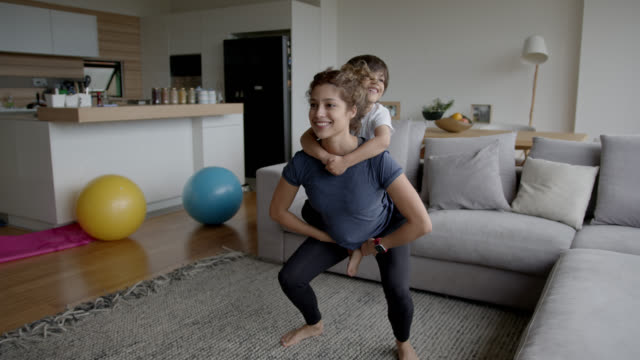 Young mother exercising at home carrying her son while doing squats both smiling