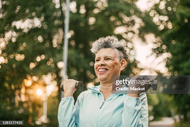 smiling wrinkled woman with dumbbells exercising in park - fitness or vitality or sport and women stockfoto's en -beelden