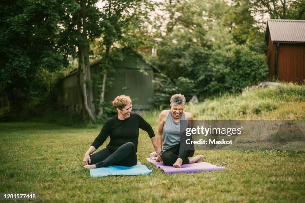 smiling woman assisting friend while exercising on mat in public park - yoga frau stock-fotos und bilder