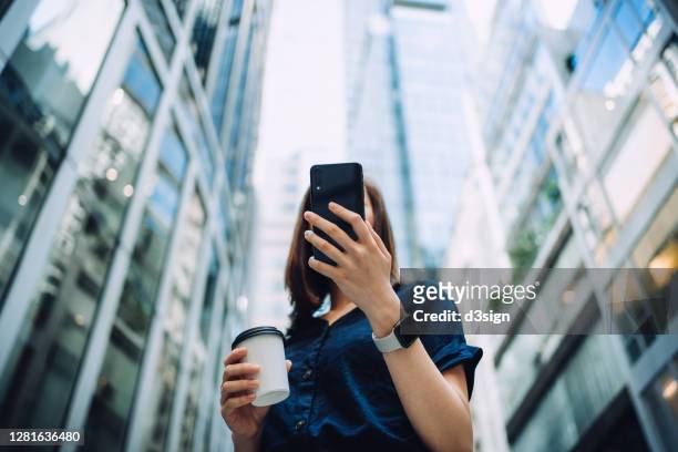 low angle portrait of confidence and successful young asian businesswoman with coffee to go, using smartphone while standing against highrise corporate buildings in financial district in the city - banking stock pictures, royalty-free photos & images