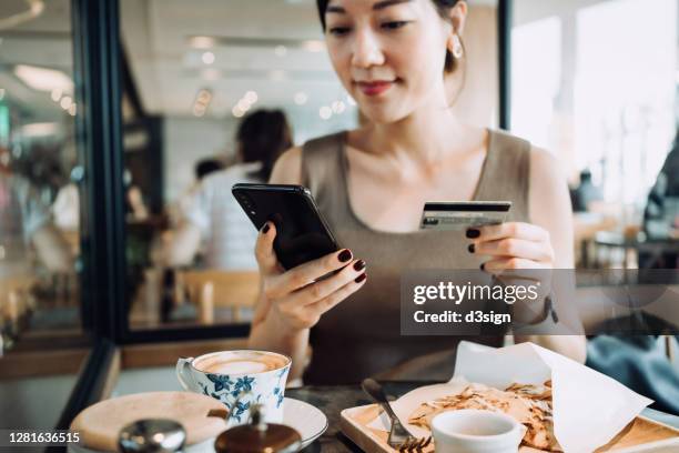 beautiful smiling young asian woman managing online banking with smartphone and making mobile payment with credit card on hand while having meal in a restaurant - spending money stock pictures, royalty-free photos & images