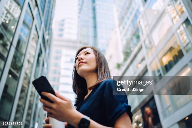 low angle portrait of confidence and successful young asian businesswoman with coffee to go, using smartphone while standing against highrise corporate buildings in financial district in the city - low motivation stock pictures, royalty-free photos & images