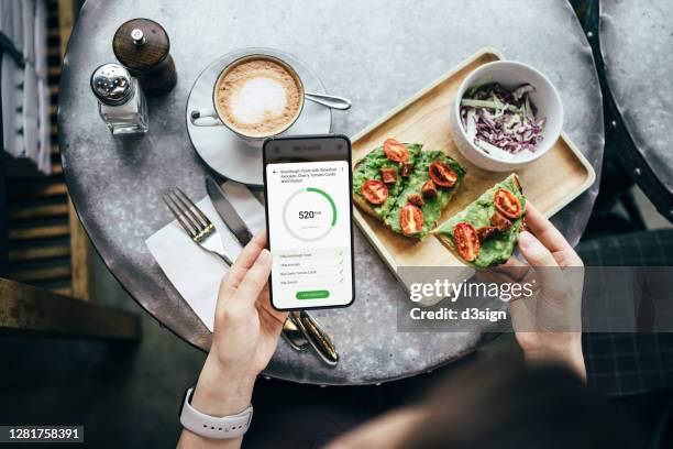 overhead view of young asian woman using fitness plan mobile app on smartphone to tailor make her daily diet meal plan, checking the nutrition facts and calories intake of her meal, sourdough toast with smashed avocado and cherry tomatoes in a restaurant - nutricionista imagens e fotografias de stock