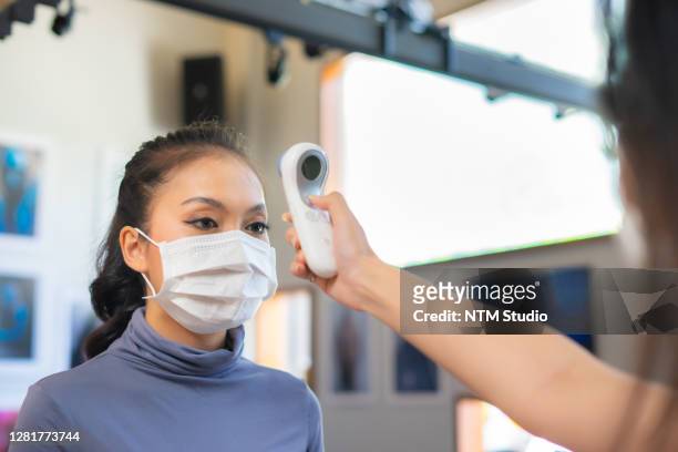 staff using non-contact infrared forehead thermometer gun to scan  body temperature of asian female customer who wearing surgical face mask during new normal measure due to coronavirus pandemic. - digital thermometer stock pictures, royalty-free photos & images