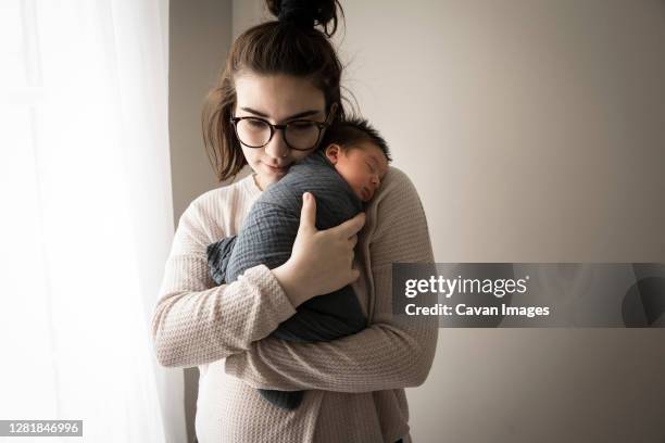hipster millennial mom snuggles swaddled newborn son next to window - baby blanket stock pictures, royalty-free photos & images