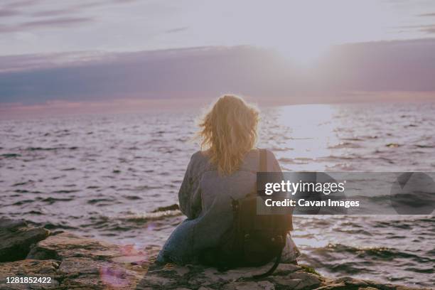 back view of blond woman sitting by the sea at sunset - bagpack stock pictures, royalty-free photos & images