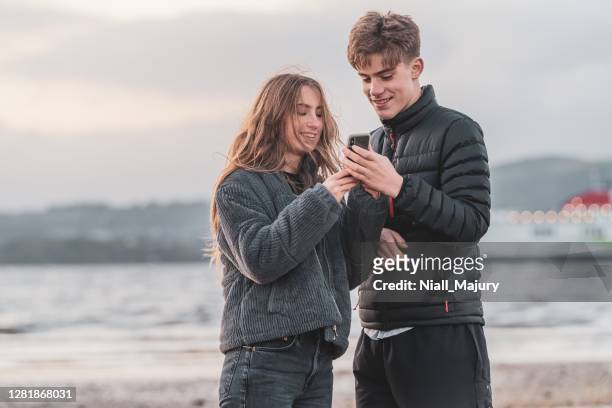 teenagers on a beach looking at a mobile phone - northern ireland coast stock pictures, royalty-free photos & images