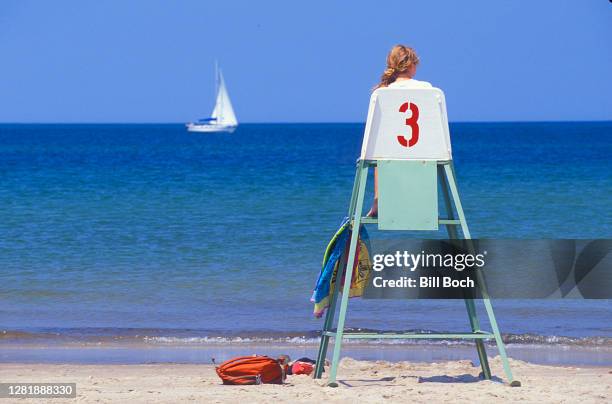 female lifeguard at a beach watching from a high lifeguard chair as a white sailboat passes in the background - beach lifeguard stock pictures, royalty-free photos & images