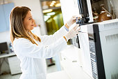 Female scientist in a white lab coat putting vial with a sample for an analysis on a gas chromatograph in biomedical lab