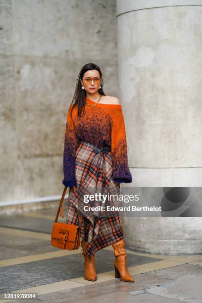 Julia Comil wears orange clear sunglasses, pearl earrings, a golden necklace, a one shoulder oversized wool orange and blue pullover with flared long...