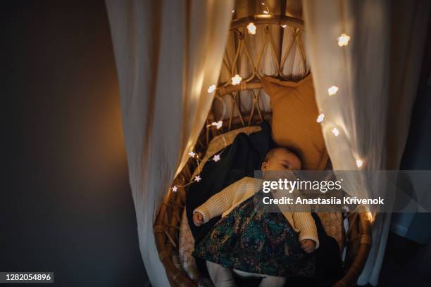 sleepy boy taking a nap inside of cozy crib with canopy and garland at home. high angle view of baby sleeping with illuminated string lights on rattan moses basket. christmas decoration around. - nursery night stock pictures, royalty-free photos & images