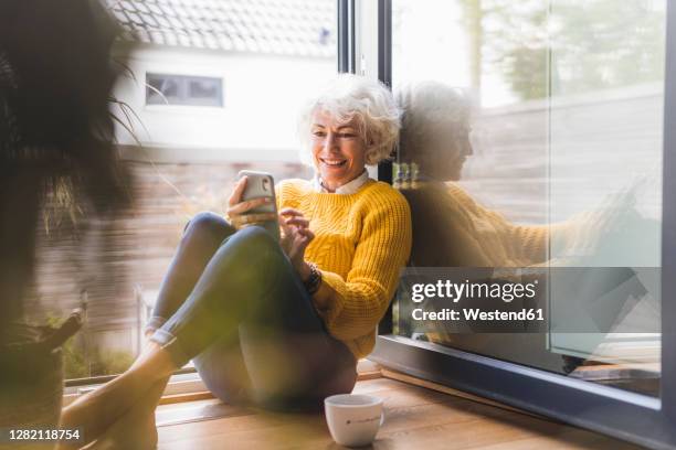 smiling woman text messaging on smart phone while sitting at home - frau zwischen 50 und 60 stock-fotos und bilder