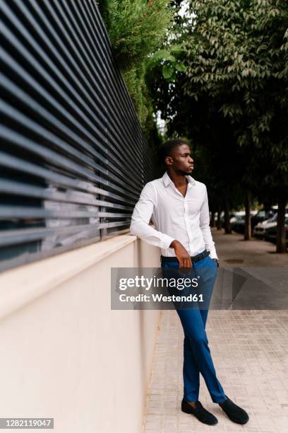 young male entrepreneur looking away while leaning on surrounding wall in city - surrounding wall stock-fotos und bilder