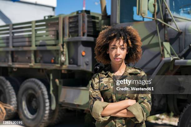 confident young female military soldier standing with arms crossed against truck at base on sunny day - military base foto e immagini stock