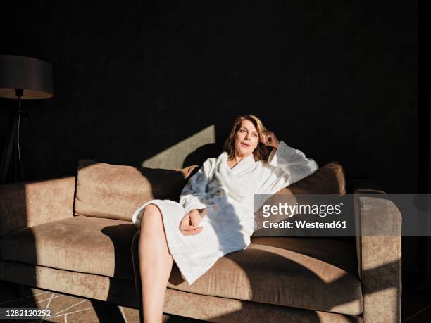 thoughtful senior woman looking away while relaxing on sofa in hotel - woman in bathrobe stock pictures, royalty-free photos & images
