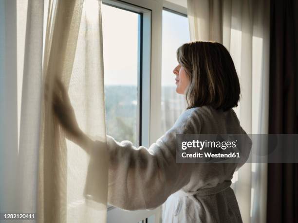 blond senior woman opening white curtain while looking through window at luxury hotel room - curtain hotel stock-fotos und bilder