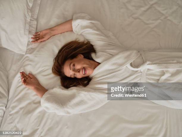 smiling senior woman lying on white bed at luxury hotel room - woman in bathrobe stock pictures, royalty-free photos & images