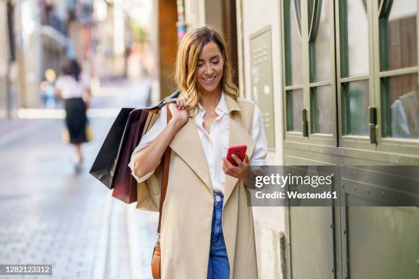 woman with shopping bag using mobile phone while standing at footpath in city - bolsa de papel fotografías e im