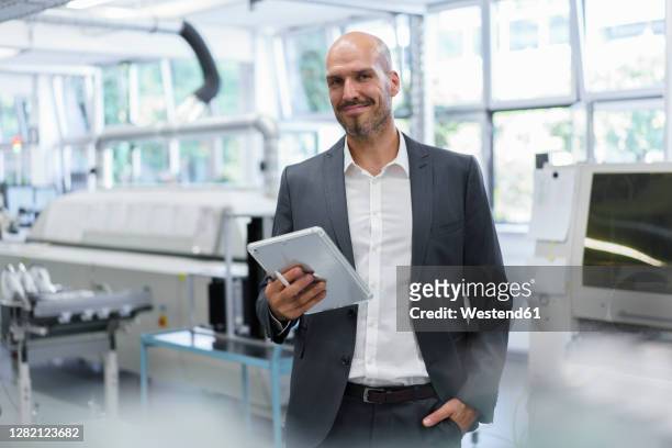 smiling confident businessman holding digital tablet while standing with hand in pocket at bright factory - three quarter length stock pictures, royalty-free photos & images